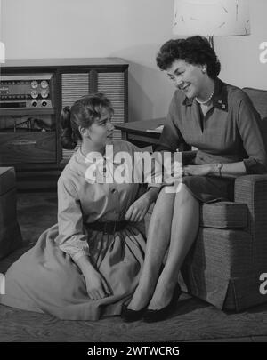 Well-dressed woman sitting in a chair in the living room. Her teenage daughter in a poodle skirt sits on the floor in front of her as they look at each other smiling Stock Photo