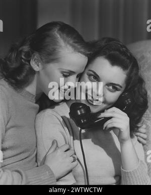 Two young teenage girls in woolen sweaters smiling, one of them speaks on the telephone as the other listens in Stock Photo