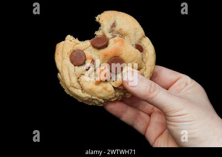 A hand holding a chocolate chip cookie with a bite taken out of it on a black background Stock Photo