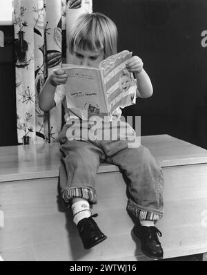 Little Boy in jeans and a bowl haircut sitting on a wooden chest reading a book titled the 'First Year of Life' Stock Photo