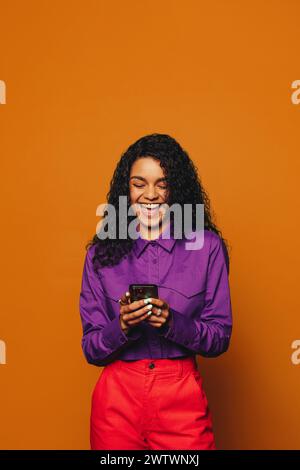 Cheerful woman with curly hair stands in a vibrant studio. She uses a mobile phone for messaging and social media, showcasing a fun, modern lifestyle. Stock Photo