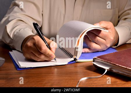 In the picture, a man who is sitting at a table holds a page of a notebook with one hand, and writes something with the other. Stock Photo