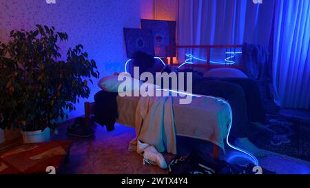 Worried, tired man sitting on bed at night and listening to music. Media. Young man can't sleep at night in a room with neon lighting. Stock Photo