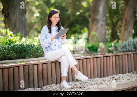 Engrossed young female student sitting cross-legged on a park bench, deeply focused on her tablet Stock Photo