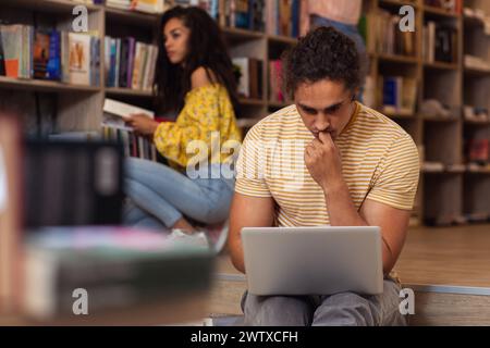 Portrait of young male student sitting at library using laptop with his friends in background. Stock Photo