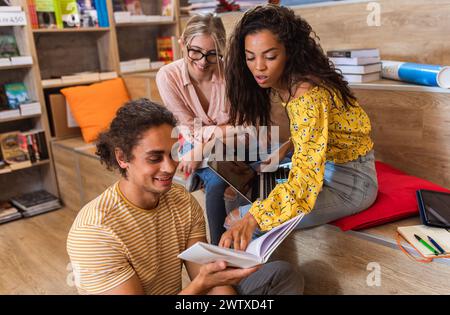 Three young students sitting on the floor in the library learning together from books and using laptop for online research. Stock Photo