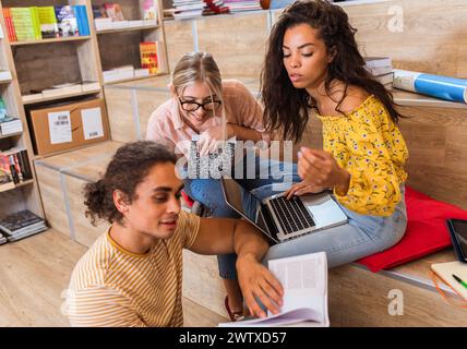 Three young students sitting on the floor in the library learning together from books and using laptop for online research. Stock Photo