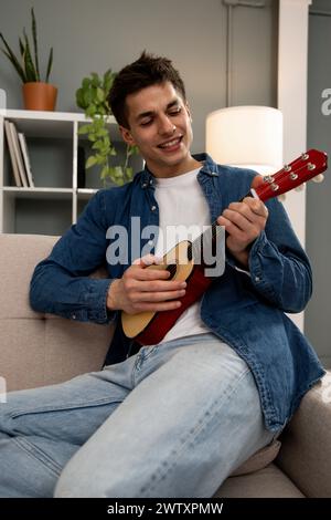 Young adult carefree male learning to play guitar on couch at living room. Young relaxed man playing music with ukelele sitting on sofa at home.  Stock Photo