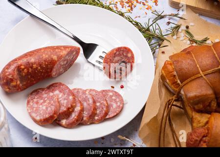 sausage cut into slices with cheese on a white plate, one piece is pinned on a fork. Nearby there is a smoked delicacy and spices Stock Photo