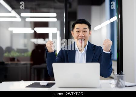An enthusiastic male professional in a suit enjoys a moment of success in front of his laptop in a modern office setting. Stock Photo
