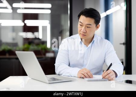 A focused businessman is engaged in work, using a laptop and making notes at his office desk in a contemporary workspace. Stock Photo
