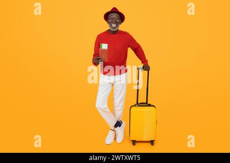 Smiling senior black man in a stylish red hat and sweater leaning on a yellow suitcase Stock Photo