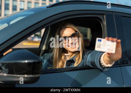 smiling young woman showing her new driver license out of car window after successful test at driving school Stock Photo