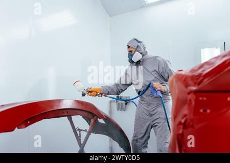 Authentic shot. Car painter in protective clothes and mask painting automobile bumper with metallic paint and varnish in chamber workshop. Stock Photo