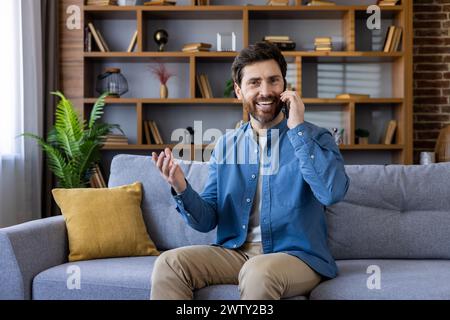 Cheerful bearded man enjoying a conversation on a mobile phone, comfortably seated on a couch in a modern living room. Stock Photo