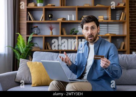 Shocked young male sitting on couch holding a credit card and using a laptop with an astonished expression. Stock Photo