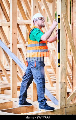 Carpenter constructing two-storey wooden frame house. Man inspects walls for levelness using spirit level, wearing protective overalls, helmet and vest. Concept of modern ecological construction. Stock Photo