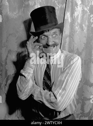 Older man in shirt and tie wearing a top hat makes a funny face with bulging eyes as he twists his mustache. Circa 1950 Stock Photo