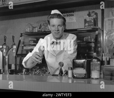 Young man in a white coat, bow tie, and a white hat, smiling at the camera while standing behind the counter of a soda shop offering up an ice cream cone and a smile Stock Photo