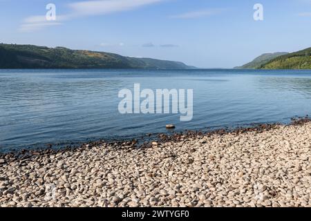 Loch Ness unfold in a carpet of smooth pebbles, transitioning to the tranquil blue waters that mirror the sky above. Surrounded by green hills Stock Photo