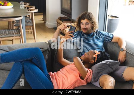A diverse couple relaxes on gray sofa at home, using a smartphone Stock Photo