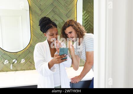 A diverse couple is examining a smartphone together while brushing teeth in a bathroom Stock Photo