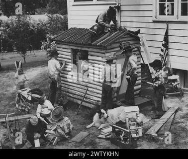 Boys in cowboy hats wearing holsters with toy guns in them building a log cabin, beside them is a baby being pushed in a carriage while a dog lays on the ground Stock Photo
