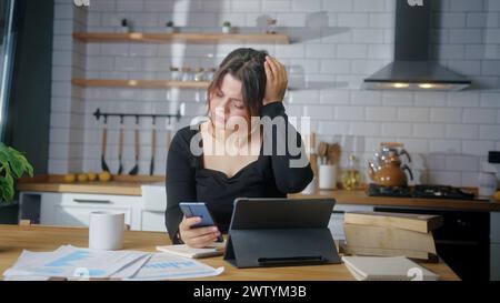 Young woman sit in the kitchen experiences severe pain headache while using smartphone in kitchen at home Stock Photo