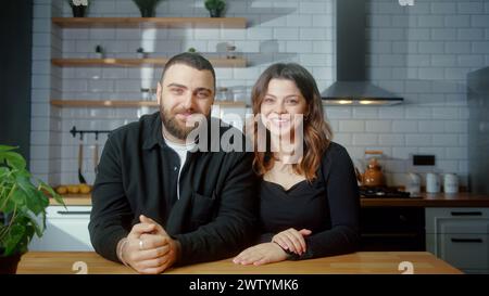Young couple sit in the kitchen make video call looking at camera wave hands greeting, talking with friends modern tech users chatting with remote dis Stock Photo