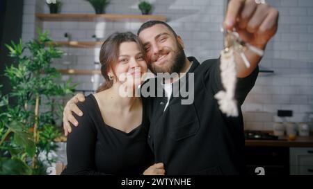 Newly married young couple sitting in kitchen smile look at camera husband show of keys from new house. Proud homeowners, rented flat, loan mortgage c Stock Photo