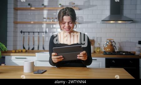 Young woman playing a video game on a tablet computer while sitting in kitchen at home. Excited concentrated woman engrossed in interesting racing gam Stock Photo