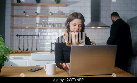 Focused representative call center agent in wireless headset helping client with complaints using laptop. Female operator working at home in kitchen, Stock Photo