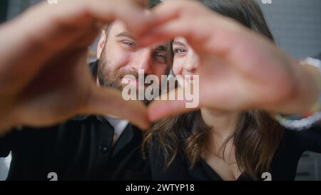 Close-up portrait of young happy couple making heart shape with joined fingers and looking at camera with smiles in between. Stock Photo