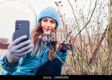 Gardener blogger filming vlog about pruning rose bush in spring garden with secateur. Woman using smartphone camera streaming on social media Stock Photo