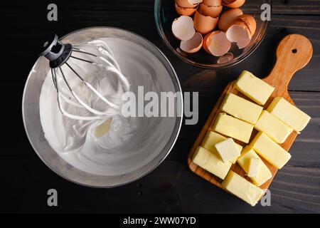 Mixing Bowl Filled with Meringue Next to Sticks of Butter: Wire whisk attachment placed in a bowl full of Swiss meringue near cracked eggs and butter Stock Photo
