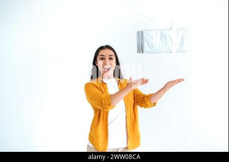 Cheerful happy brunette indian or arabian woman in casual wear, stands on isolated white background, throws up big gift box, looking at camera and smiles friendly. Gifts and surprises concept Stock Photo