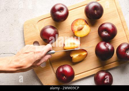 A woman's hand cutting a plum with a knife on a wooden cutting board an grey background. Stock Photo