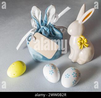 Easter image with creative gift bag with bunny ears, blank note, white ceramic bunny, decorative eggs with floral pattern on gray background. Mock up. Stock Photo