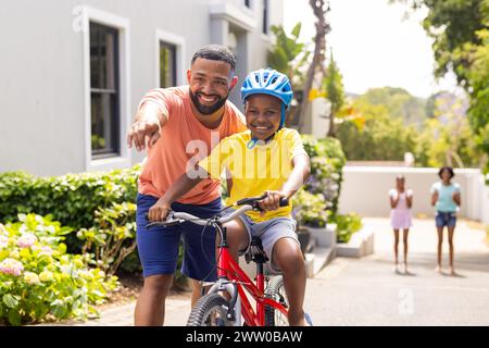 African American father teaches son to ride a bike outside home, family watches Stock Photo