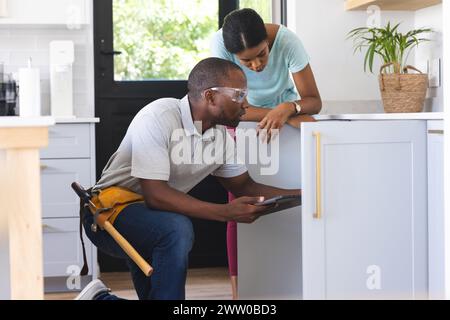 African American plumber fixing sink while using tablet while homeowner is watching Stock Photo