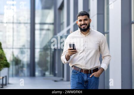 Portrait of a smiling young Nidhi man who is smiling, holding his hand in his jeans pocket, using a mobile phone and looking confidently into the camera. Stock Photo