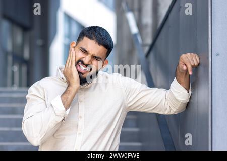 Close-up photo of a young Muslim man standing outside a building, leaning against a wall and holding his hand to his cheek, suffering from a severe toothache. Stock Photo