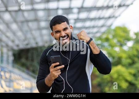 A joyful young man with headphones celebrates while holding a smartphone outside, expressing happiness and energy. Stock Photo