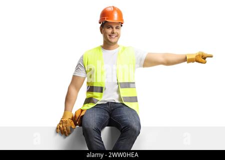 Construction worker sitting on a blank panel and pointing to the side isolated on white background Stock Photo