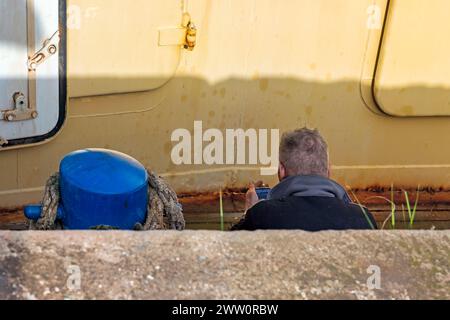 male person photographed from behind is looking at his cell phone with a cigarette in his hand sitting on the deck of a ship Stock Photo