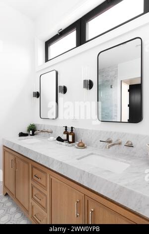 A bathroom with a white oak cabinet, marble countertop, bronze faucets, black and white tiled floor, and sconces around black mirrors. Stock Photo