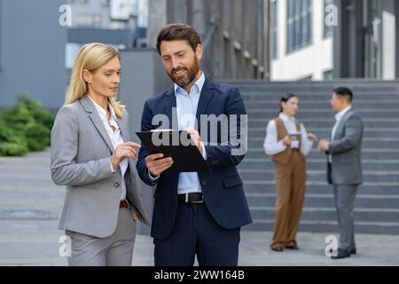 Two corporate professionals engaging in a discussion with a digital tablet, with colleagues conversing in the background. Stock Photo