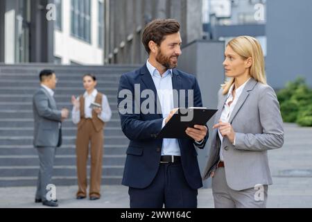 Two corporate professionals in suits engaged in a discussion with a clipboard outside a modern office building. Stock Photo