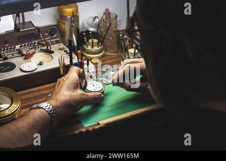 An over-the-shoulder view of a watchmaker repairing the machinery of a watch on his workbench full of various specialised tools of the watchmaking tra Stock Photo