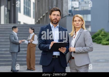 Two focused business professionals with clipboard standing outside the office, colleagues discussing in the background. Stock Photo
